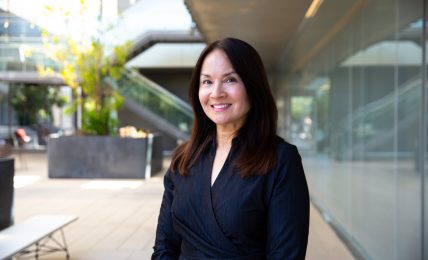 Woman with dark hair smiling, wearing a dark blazer, standing outdoors near a modern building with glass walls.