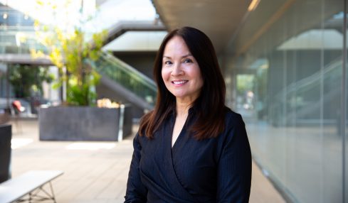 Woman with dark hair smiling, wearing a dark blazer, standing outdoors near a modern building with glass walls.