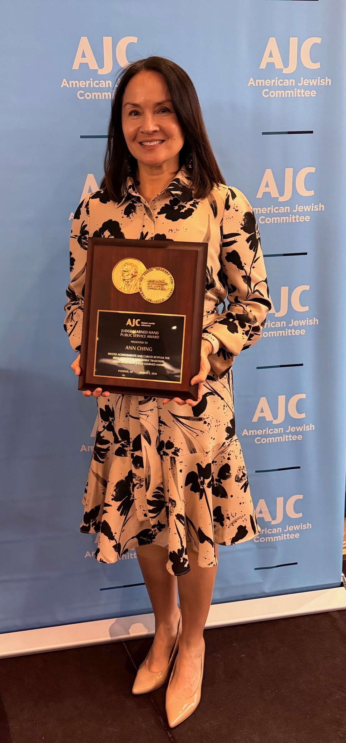 Woman holding an award plaque, smiling, standing in front of a blue AJC backdrop, wearing a floral dress and beige shoes.