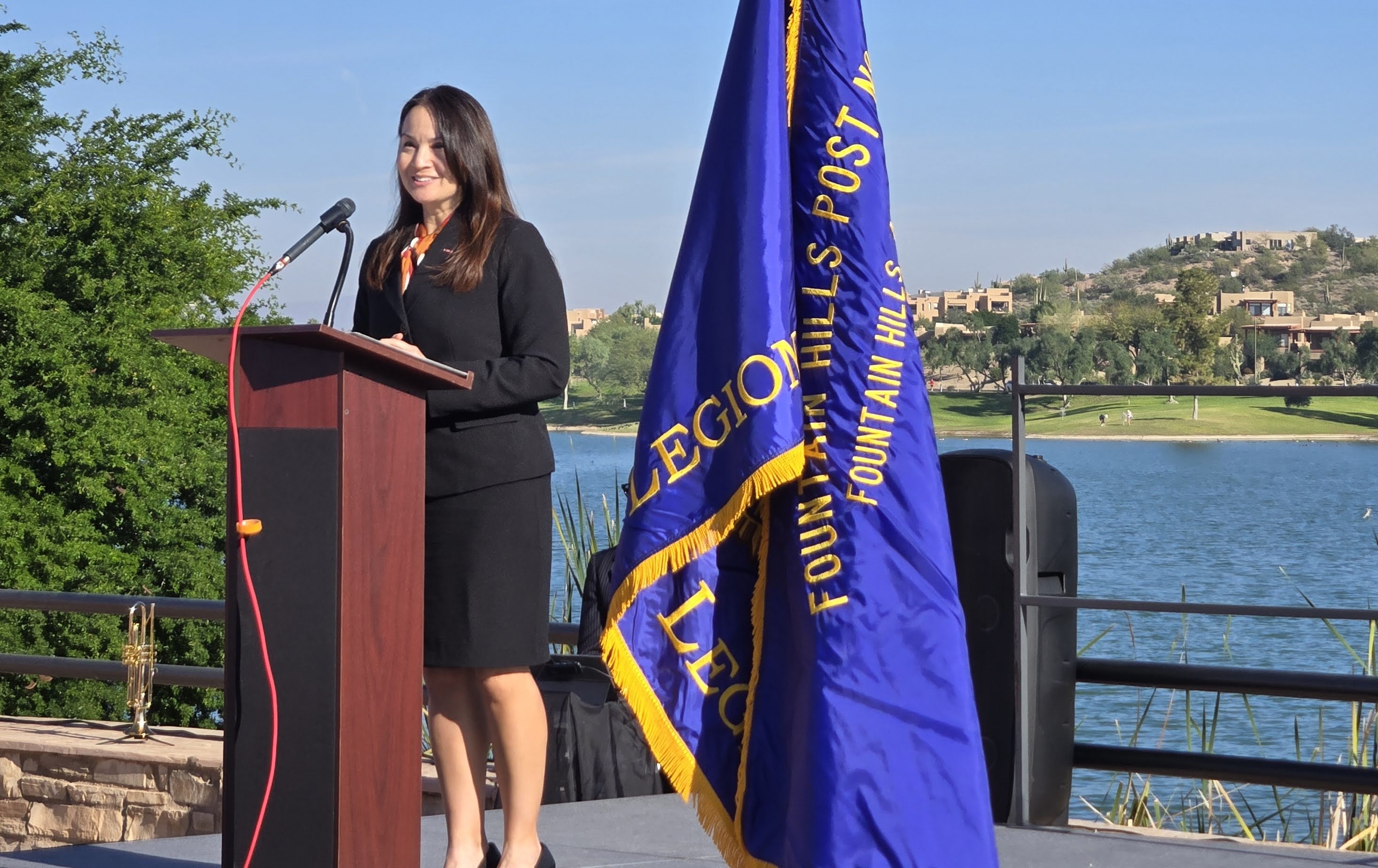 Woman in black dress speaking at podium outdoors with a blue flag and river in background.