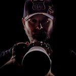Person wearing a cap, holding a camera, with a dark background, face partially illuminated.