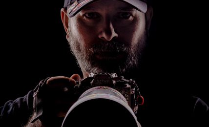 Person wearing a cap, holding a camera, with a dark background, face partially illuminated.