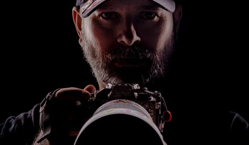 Person wearing a cap, holding a camera, with a dark background, face partially illuminated.