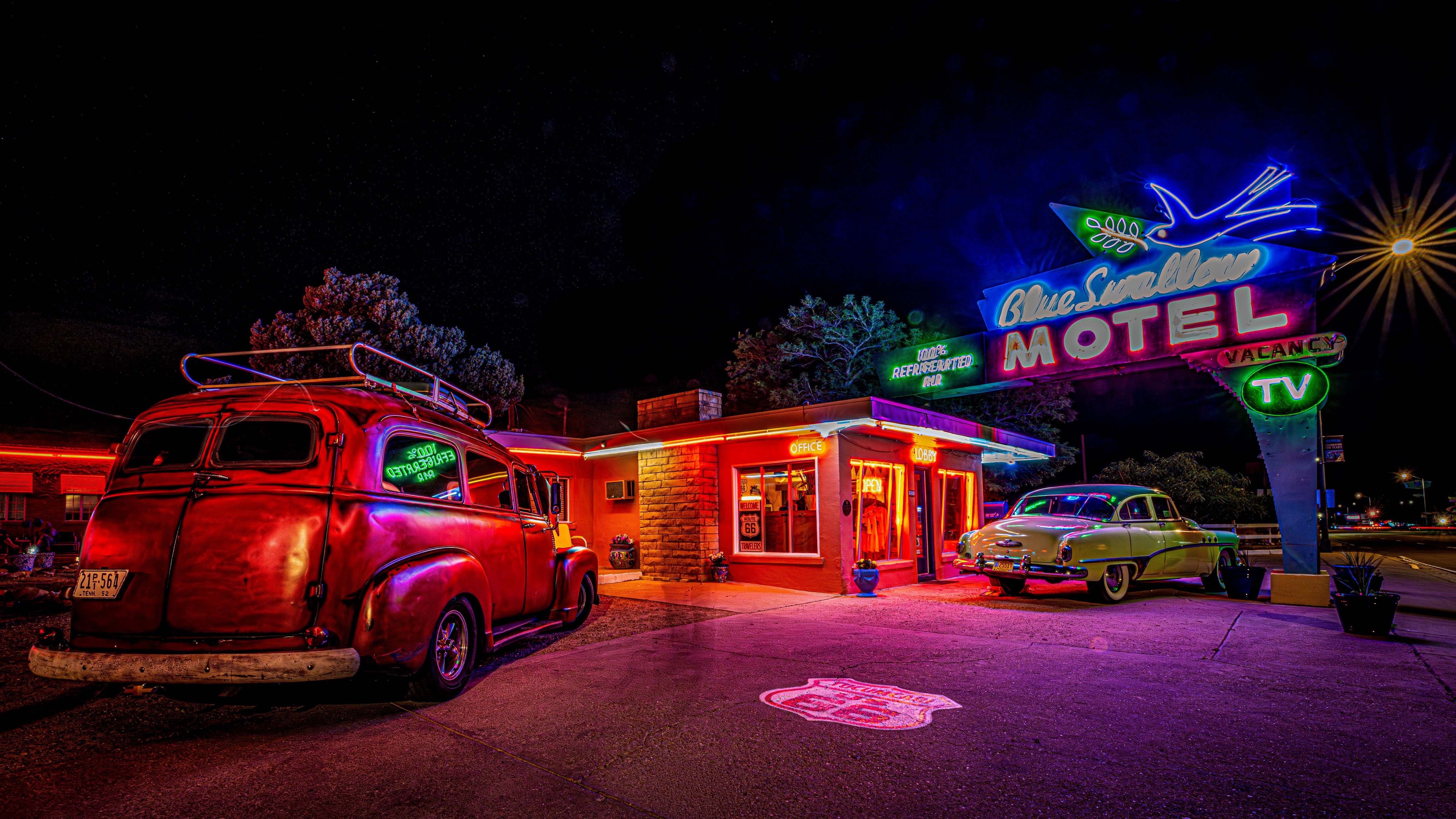 Night scene of a vintage motel with neon signs, a classic car, and a retro building illuminated in colorful lights.