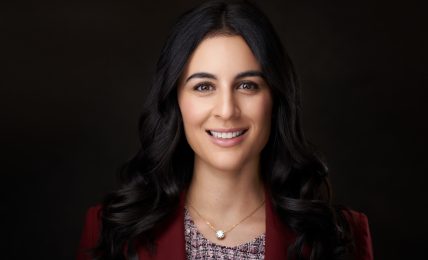 Portrait of a smiling woman with long dark hair, wearing a red blazer and a patterned top, against a dark background.