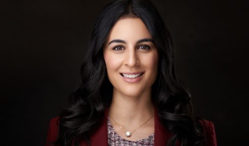 Portrait of a smiling woman with long dark hair, wearing a red blazer and a patterned top, against a dark background.