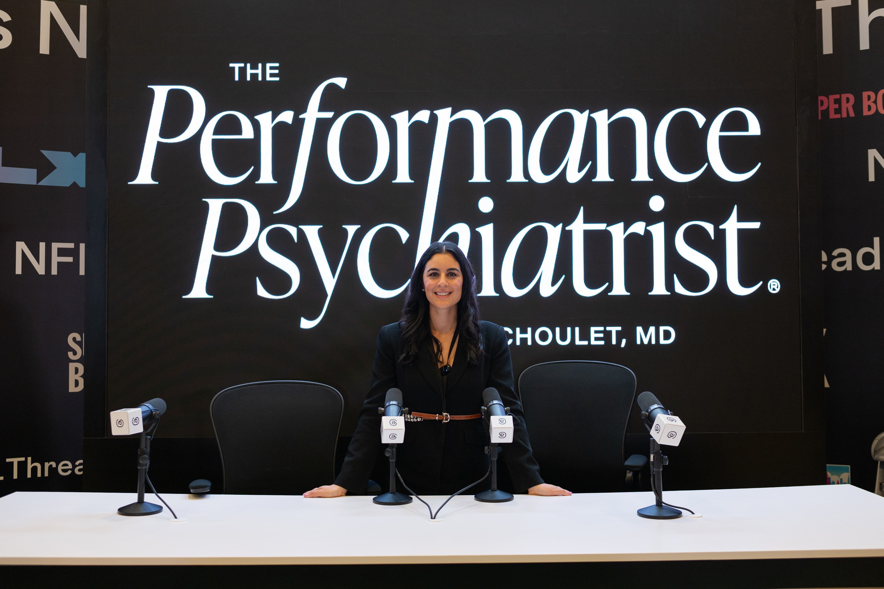 Woman sitting at a table with microphones, behind a large sign reading 'The Performance Psychiatrist, Per B. HOULET, MD'.