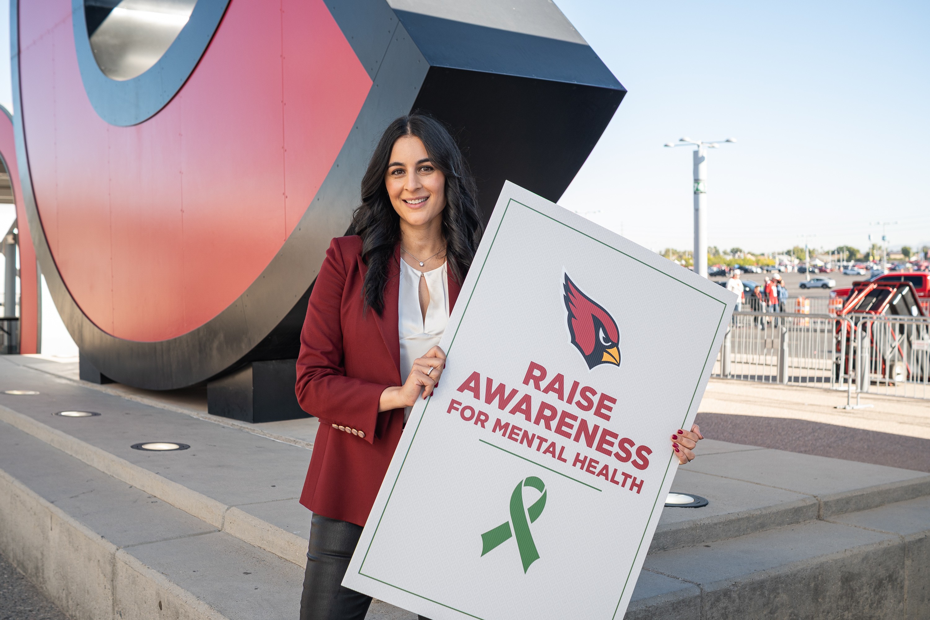 Woman holding a sign promoting mental health awareness outside a modern building, smiling, with a parking lot in background.
