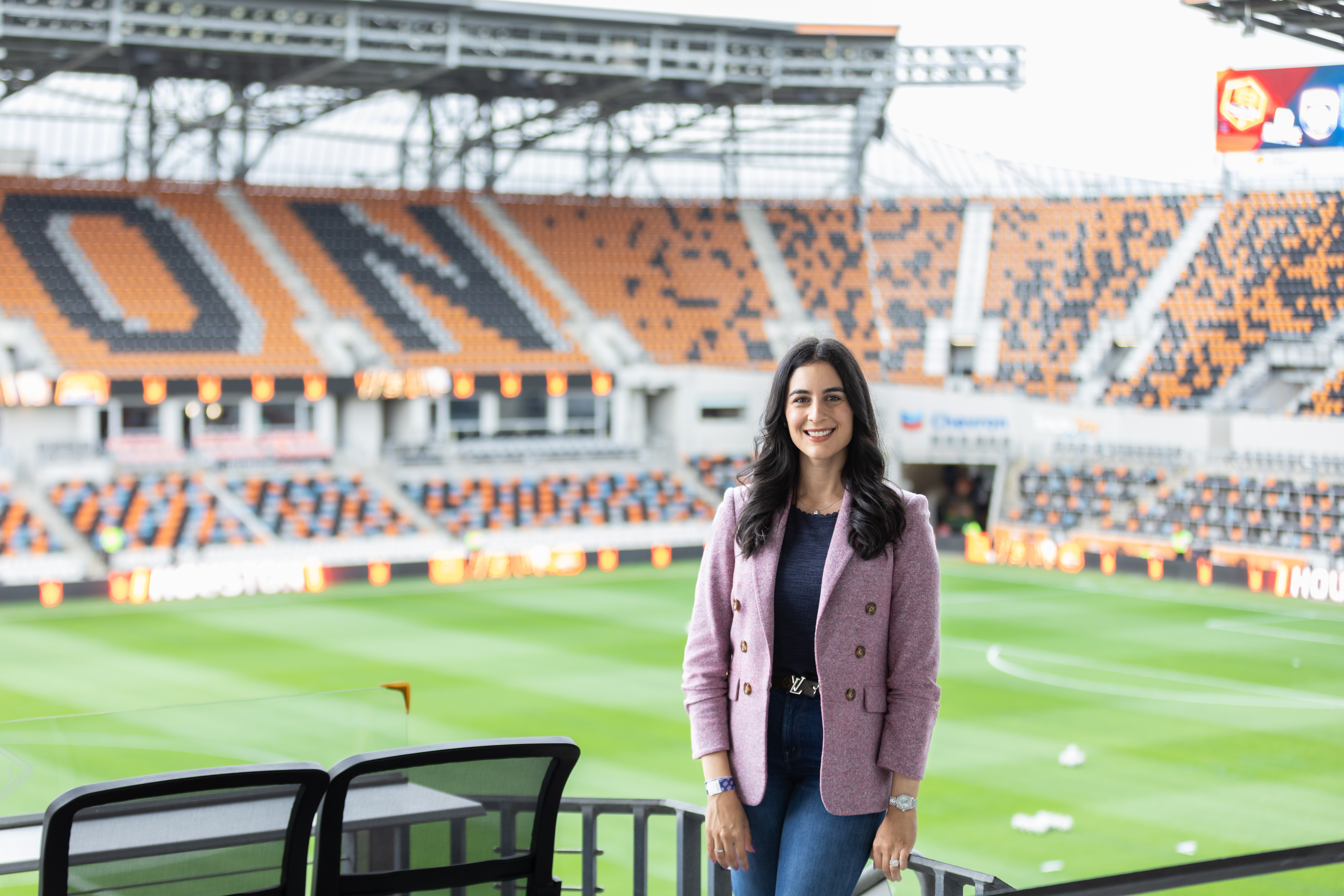 Woman standing on a sports field with stadium seating in the background, smiling, wearing a purple jacket and dark clothing.
