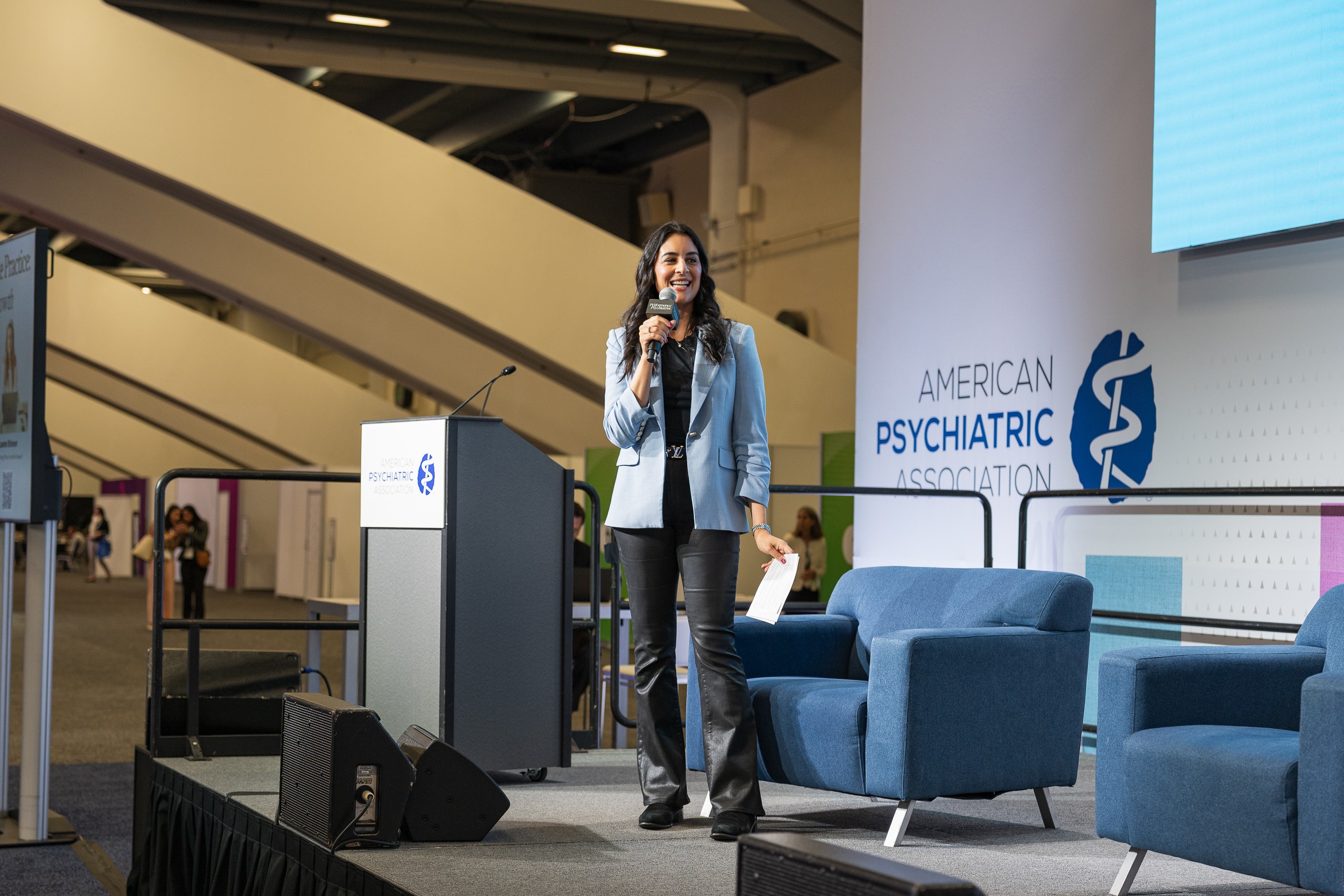 Woman speaking on stage at conference with blue chairs and a large screen, American Psychiatric Association logo visible.