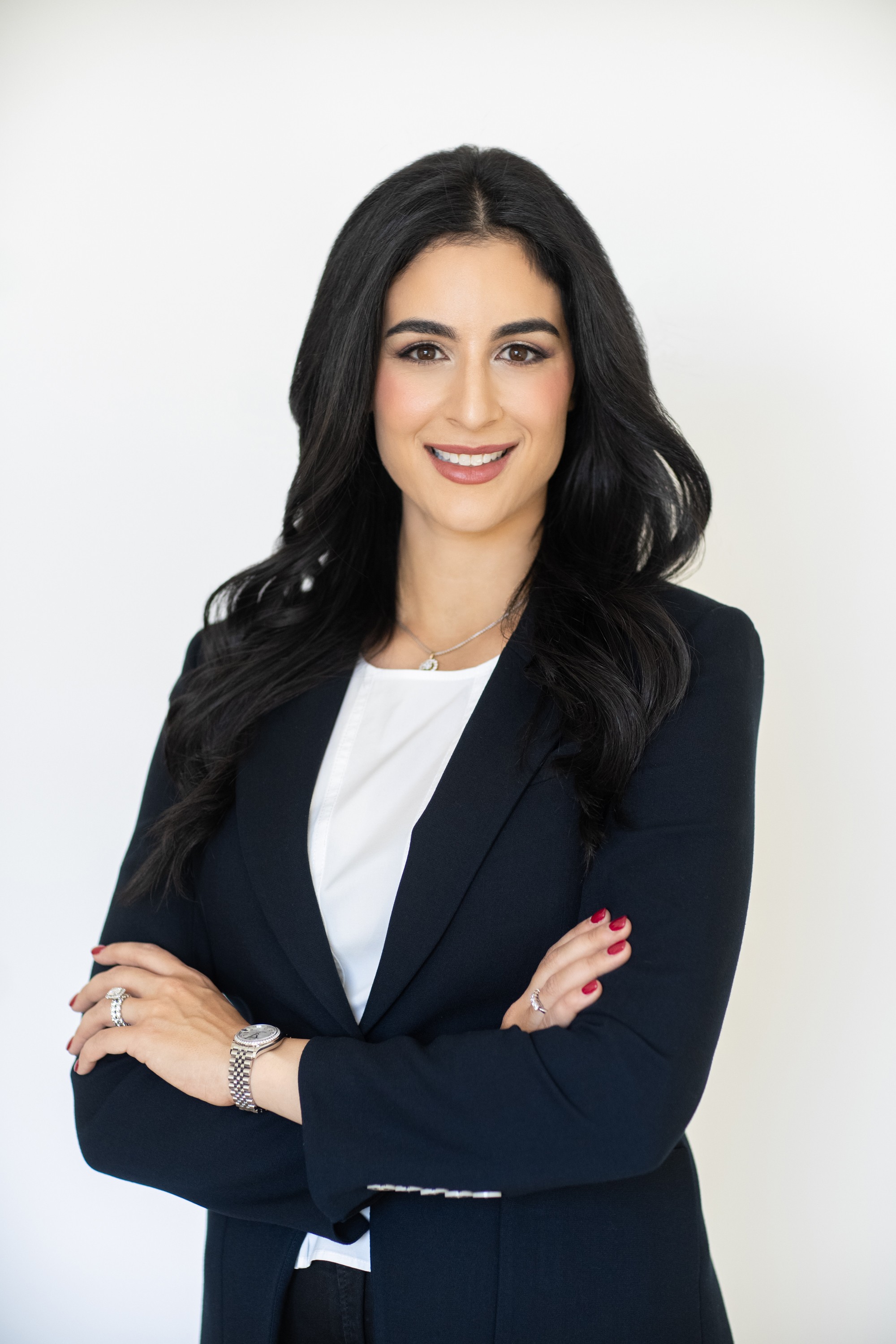 Woman with long dark hair in a navy blazer and white top, smiling with arms crossed, against a white background.