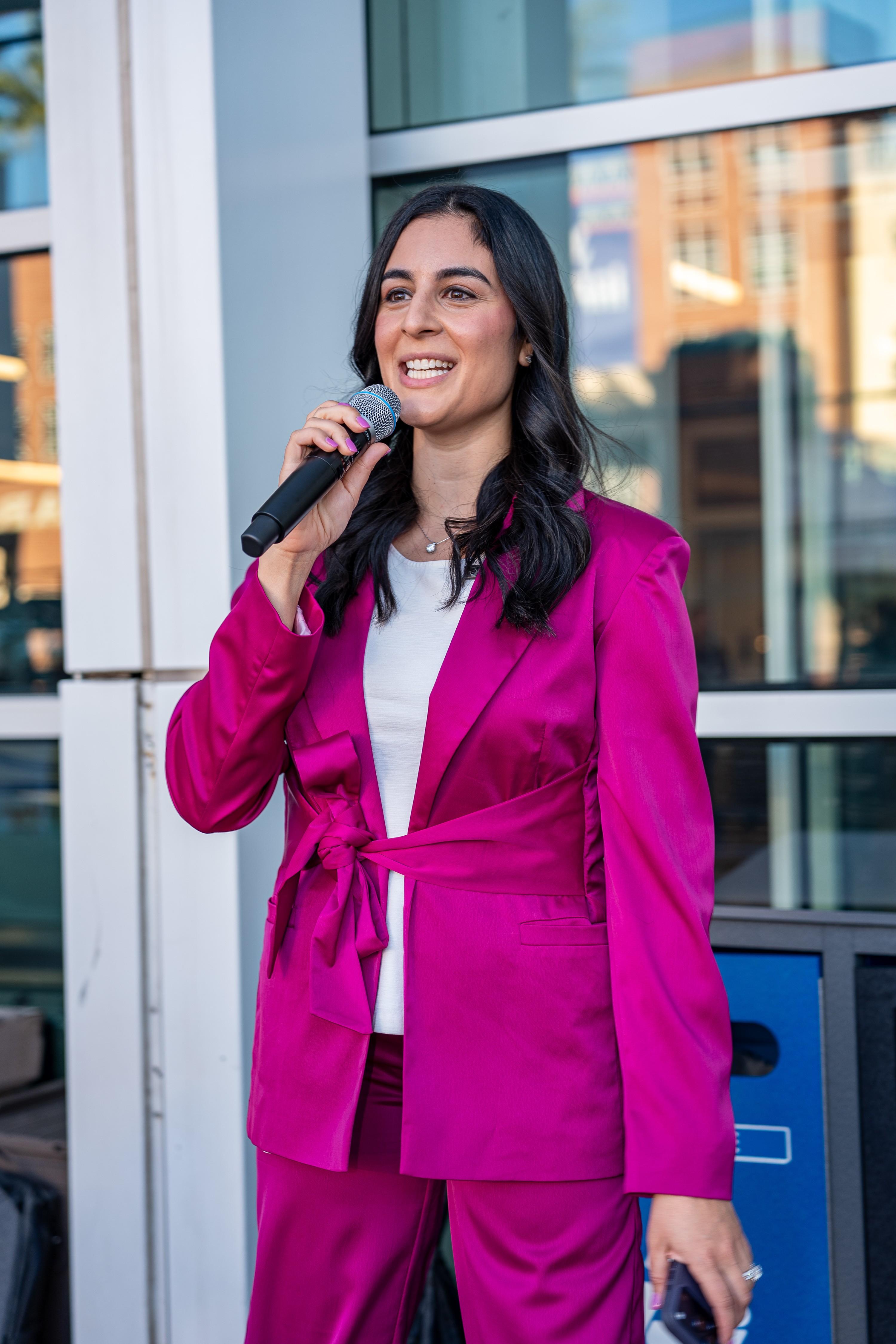 Woman in bright pink suit holding microphone, smiling outdoors with glass building background.