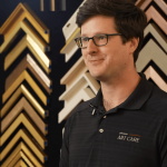 Man with glasses and dark hair wearing a black polo shirt stands in front of a display of picture frame samples.