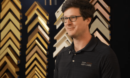 Man with glasses and dark hair wearing a black polo shirt stands in front of a display of picture frame samples.
