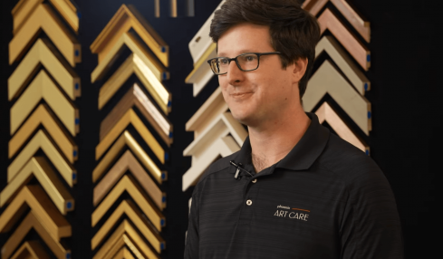 Man with glasses and dark hair wearing a black polo shirt stands in front of a display of picture frame samples.
