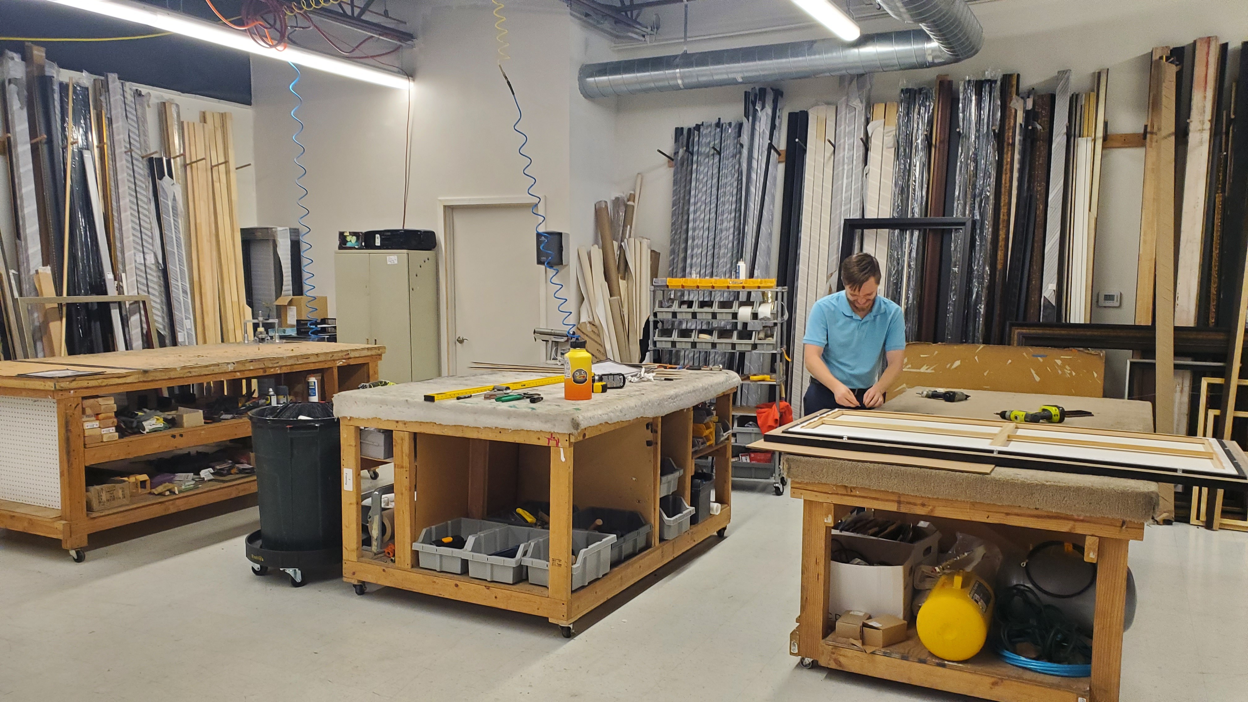 Workshop with tables, shelves, and a person working on a large wooden board, surrounded by materials and tools.