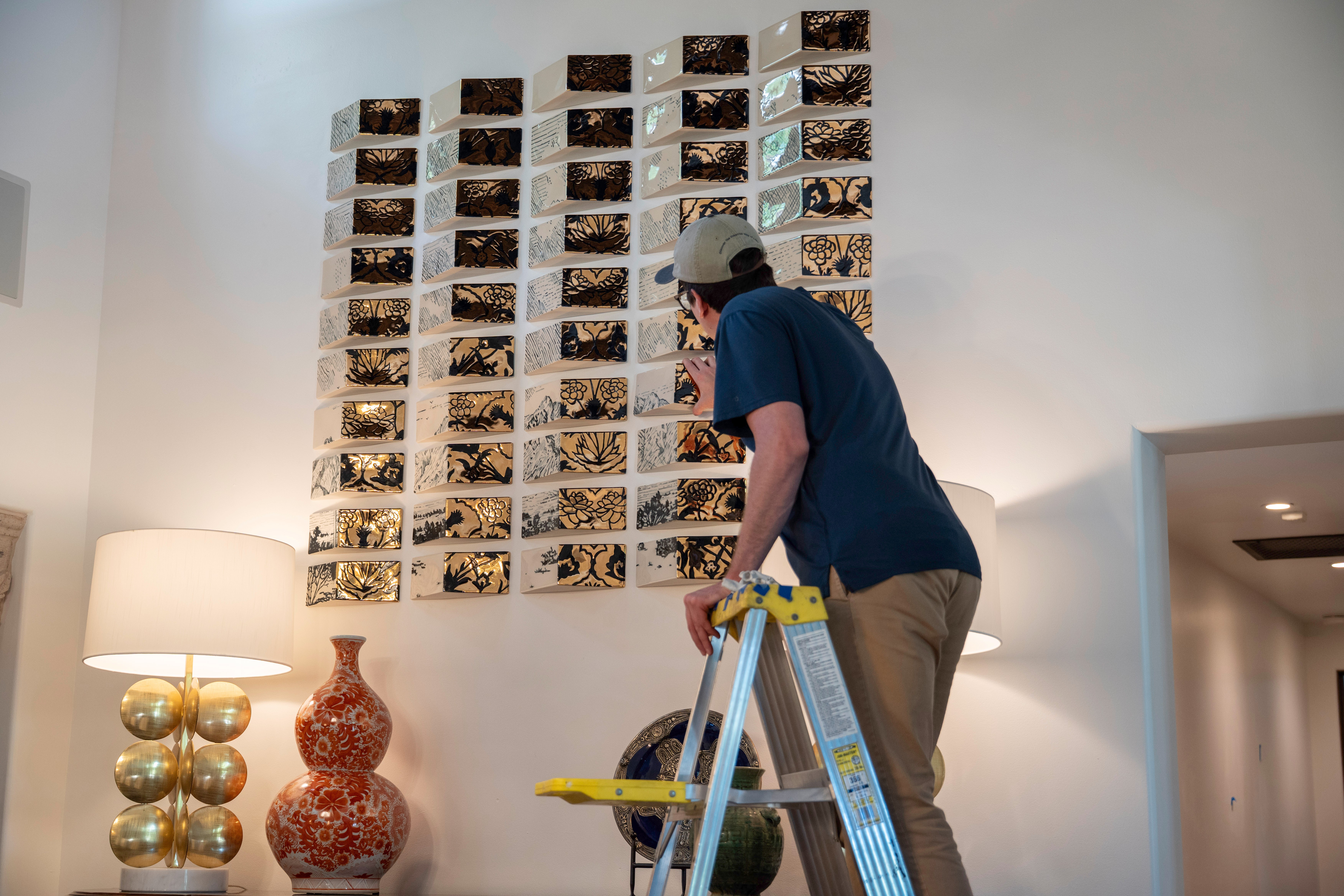 Person standing on a ladder, looking at a wall display of small photographs or images, in a room with lamps and a decorative vase.