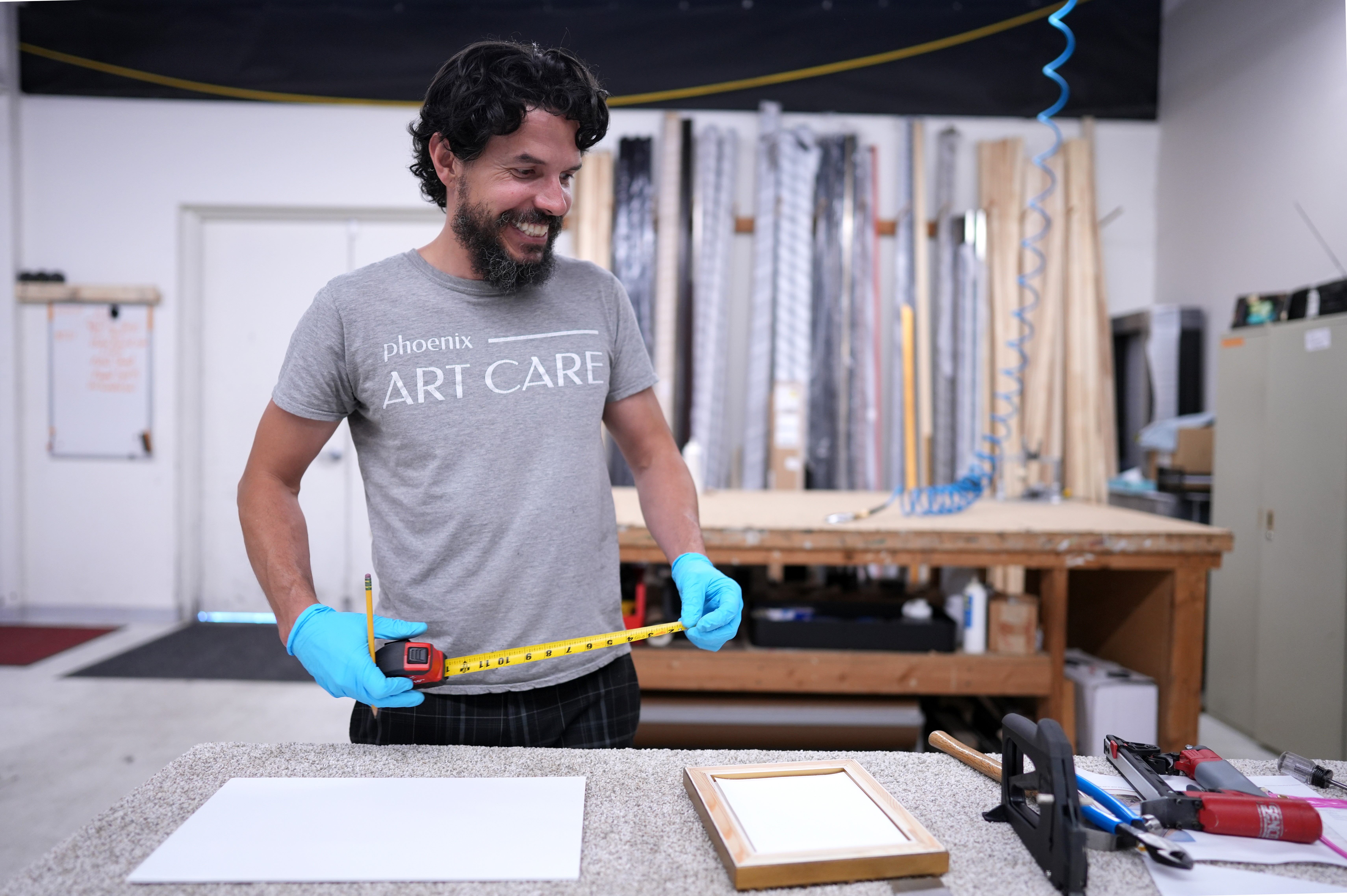 Man with dark curly hair and beard measuring a white canvas in a workshop, wearing a gray t-shirt and blue gloves.