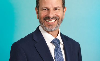 Man with a beard in a suit and tie smiling against a blue gradient background.