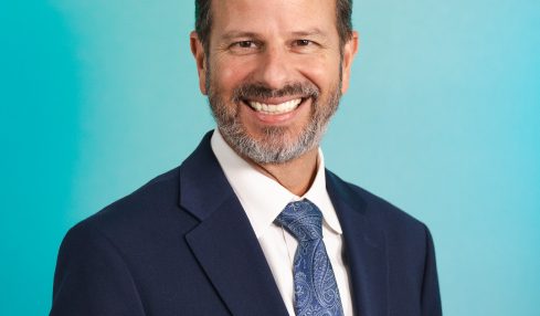 Man with a beard in a suit and tie smiling against a blue gradient background.