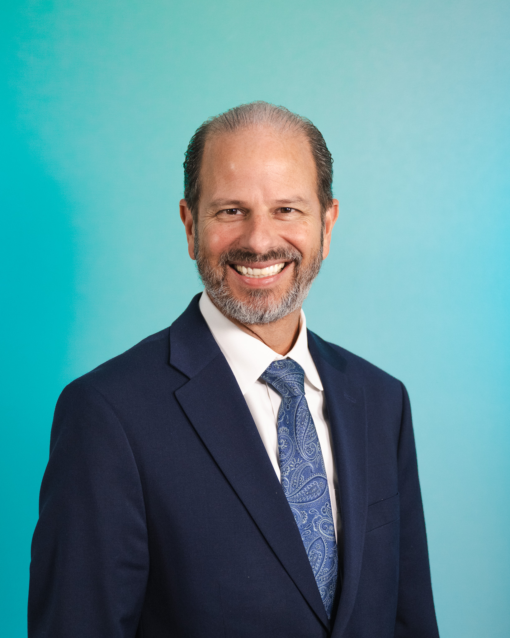 Man with a beard in a suit and tie smiling against a blue gradient background.