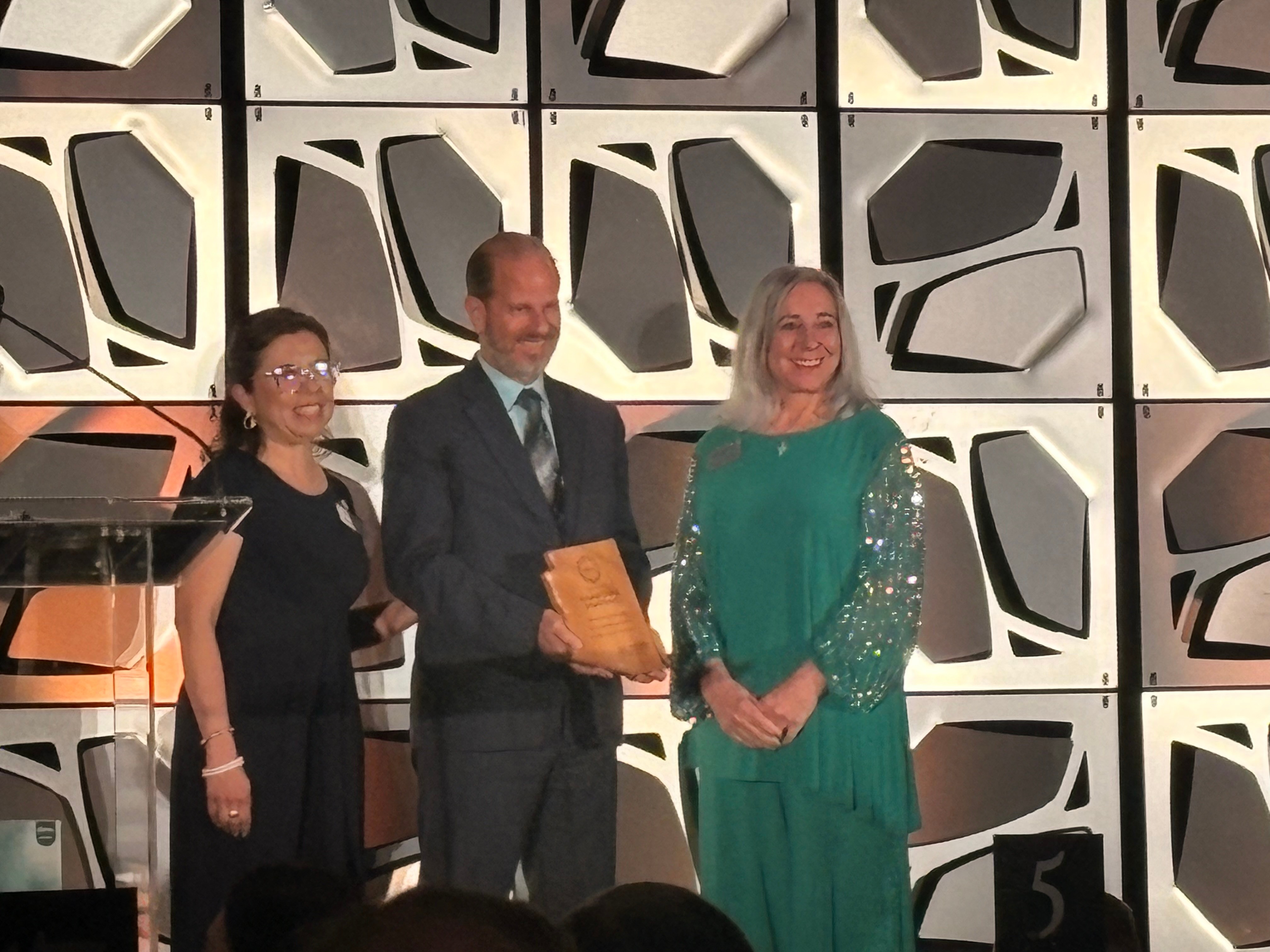 Three people standing on stage, one holding an award, with a geometric patterned backdrop.