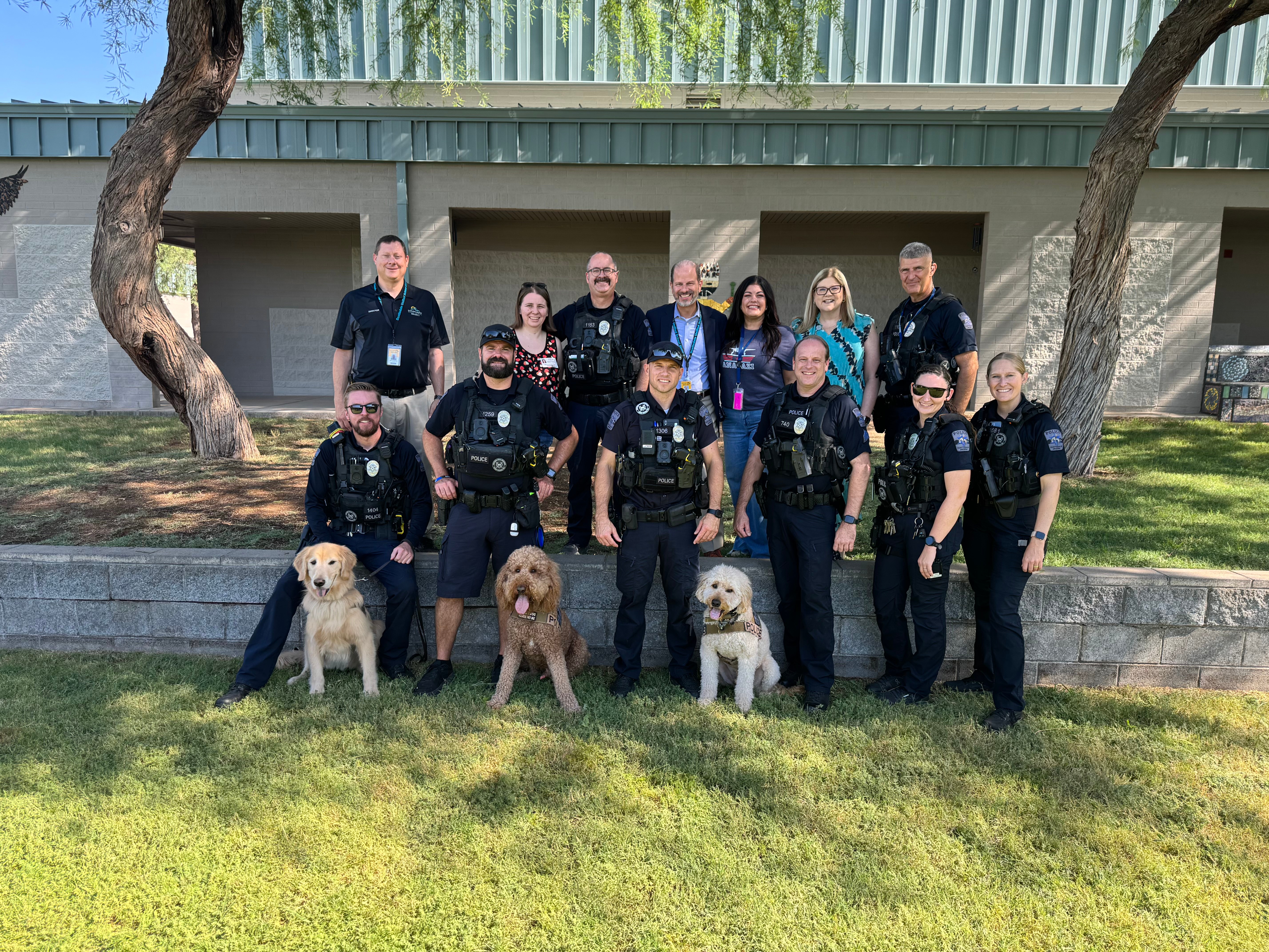 Group of police officers with four dogs standing outdoors in front of a building with trees and grass.