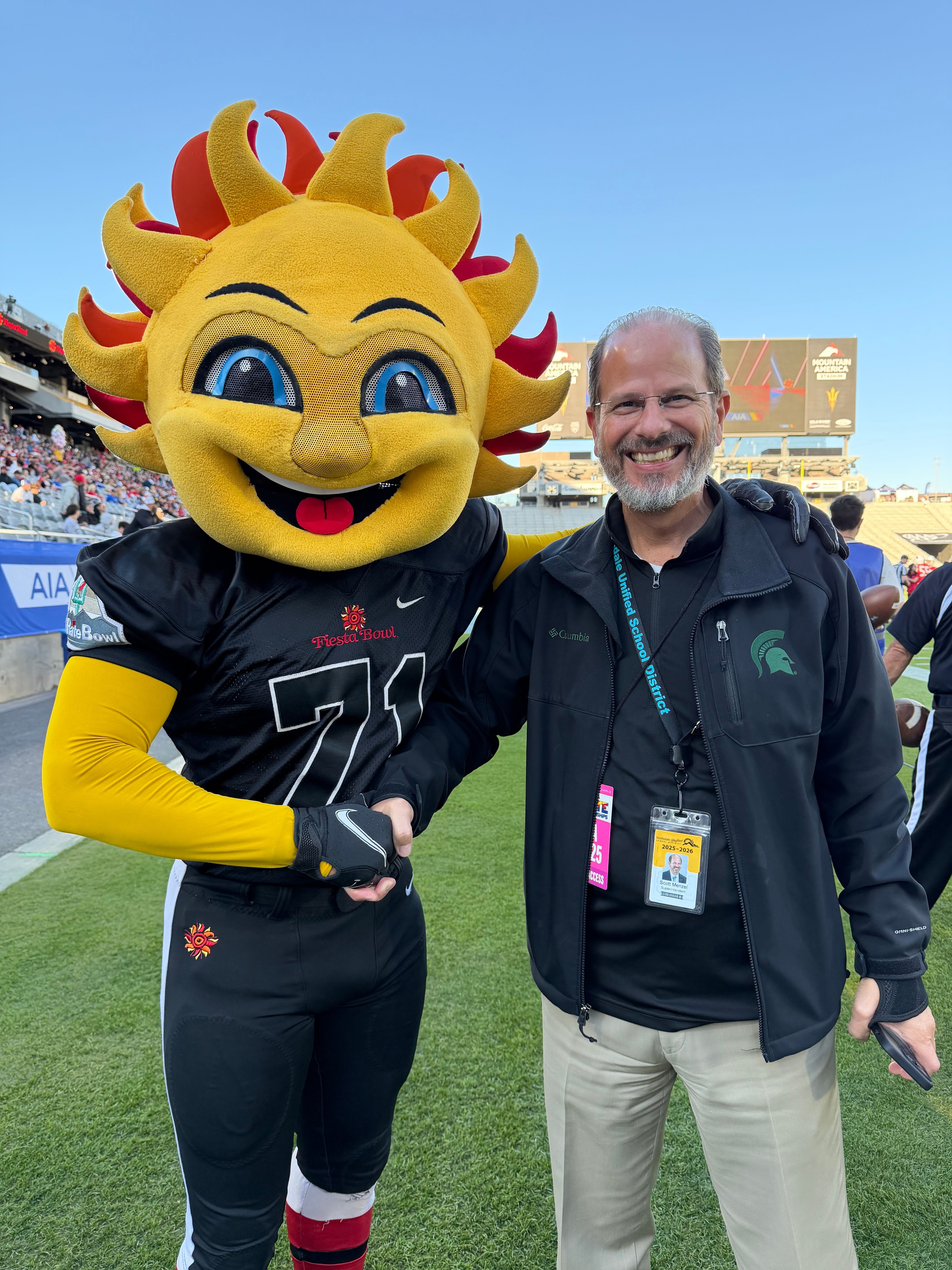 Person standing next to a mascot with a sun-like face, yellow arms, and a black sports outfit, outdoors on grass.