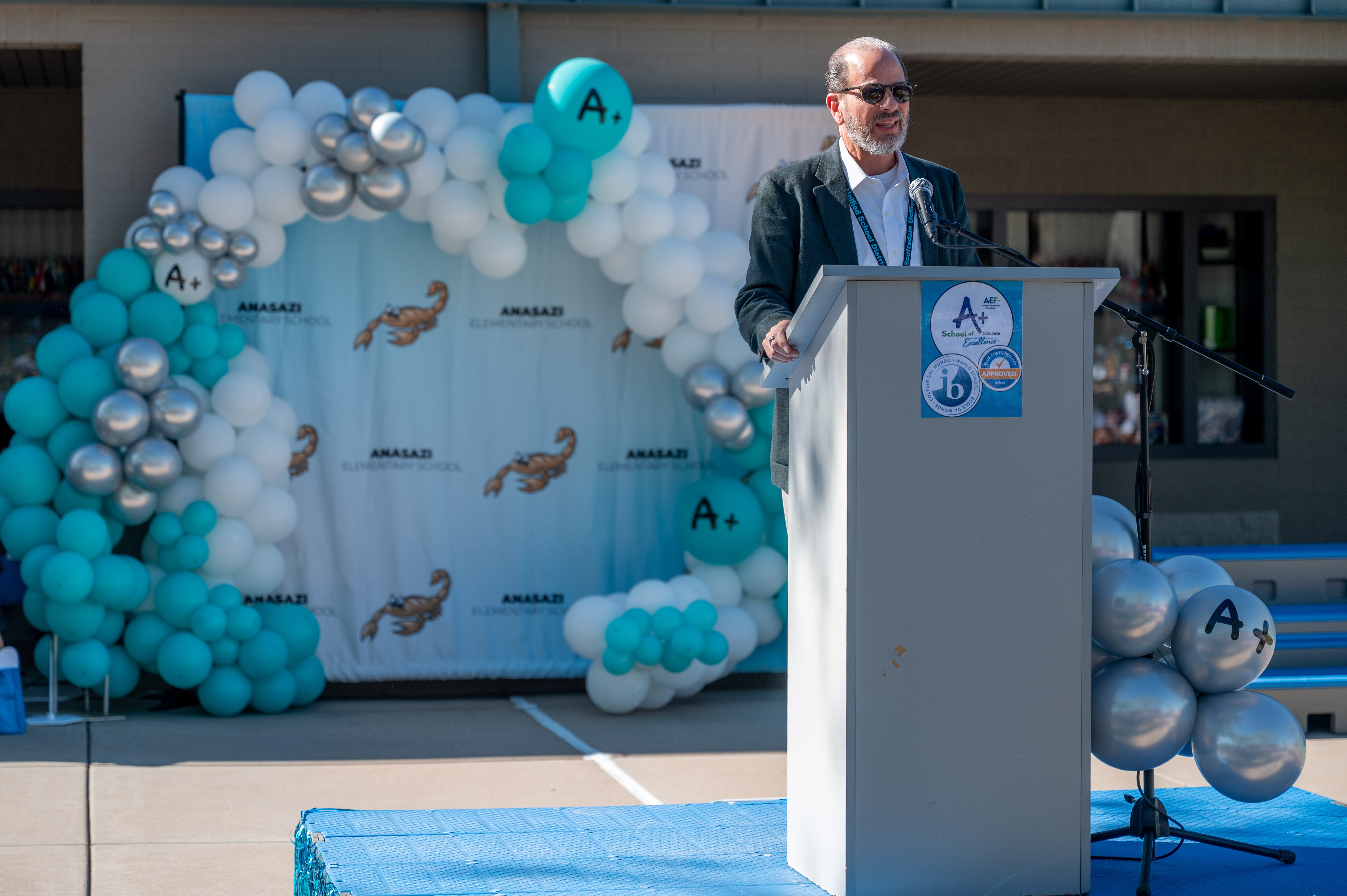 Man in suit speaking at podium with balloon decorations and backdrop featuring logos and images of animals.