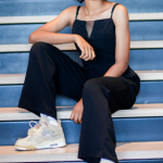Young woman with curly hair sitting on stairs, smiling, wearing a black jumpsuit and sneakers.
