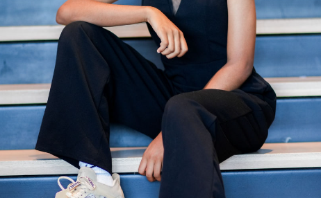 Young woman with curly hair sitting on stairs, smiling, wearing a black jumpsuit and sneakers.