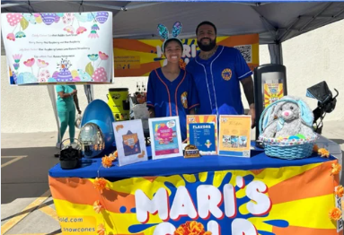 Two people stand behind a colorful table with books and a basket, under a tent at an outdoor event.