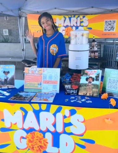 Young woman standing behind a table with colorful flyers and a large sign reading 'MARIE'S' at an outdoor event.