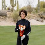 Woman with curly hair and glasses holding a golf club on a golf course with cacti in the background.
