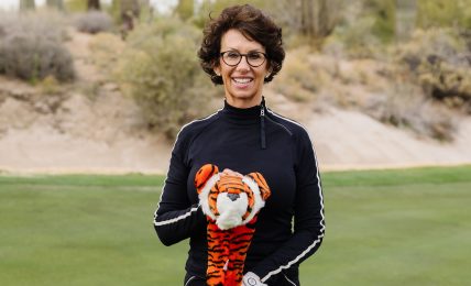 Woman with curly hair and glasses holding a golf club on a golf course with cacti in the background.