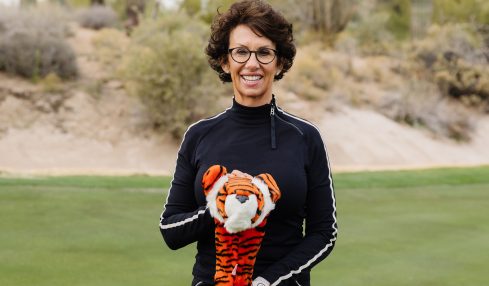Woman with curly hair and glasses holding a golf club on a golf course with cacti in the background.