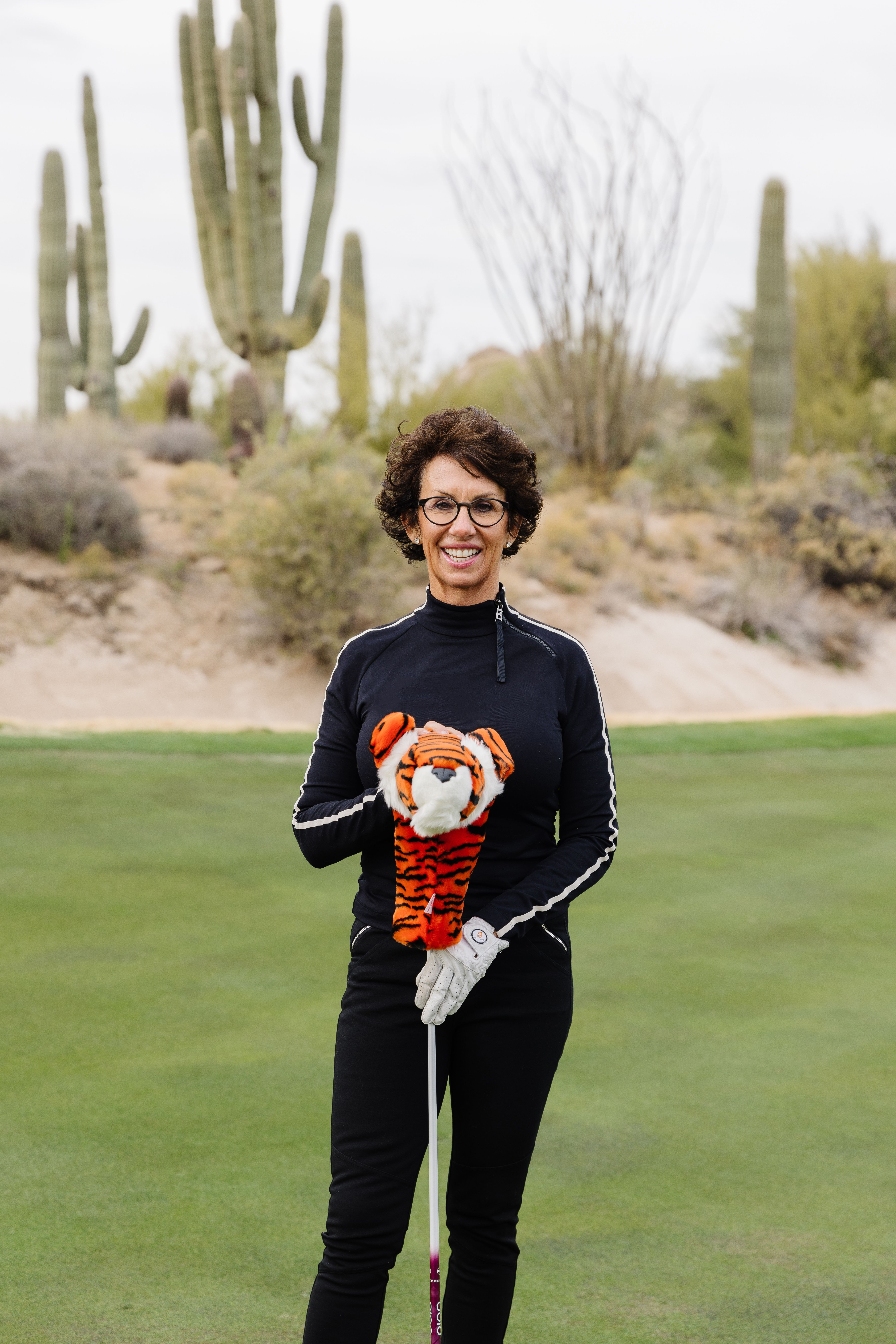 Woman with curly hair and glasses holding a golf club on a golf course with cacti in the background.