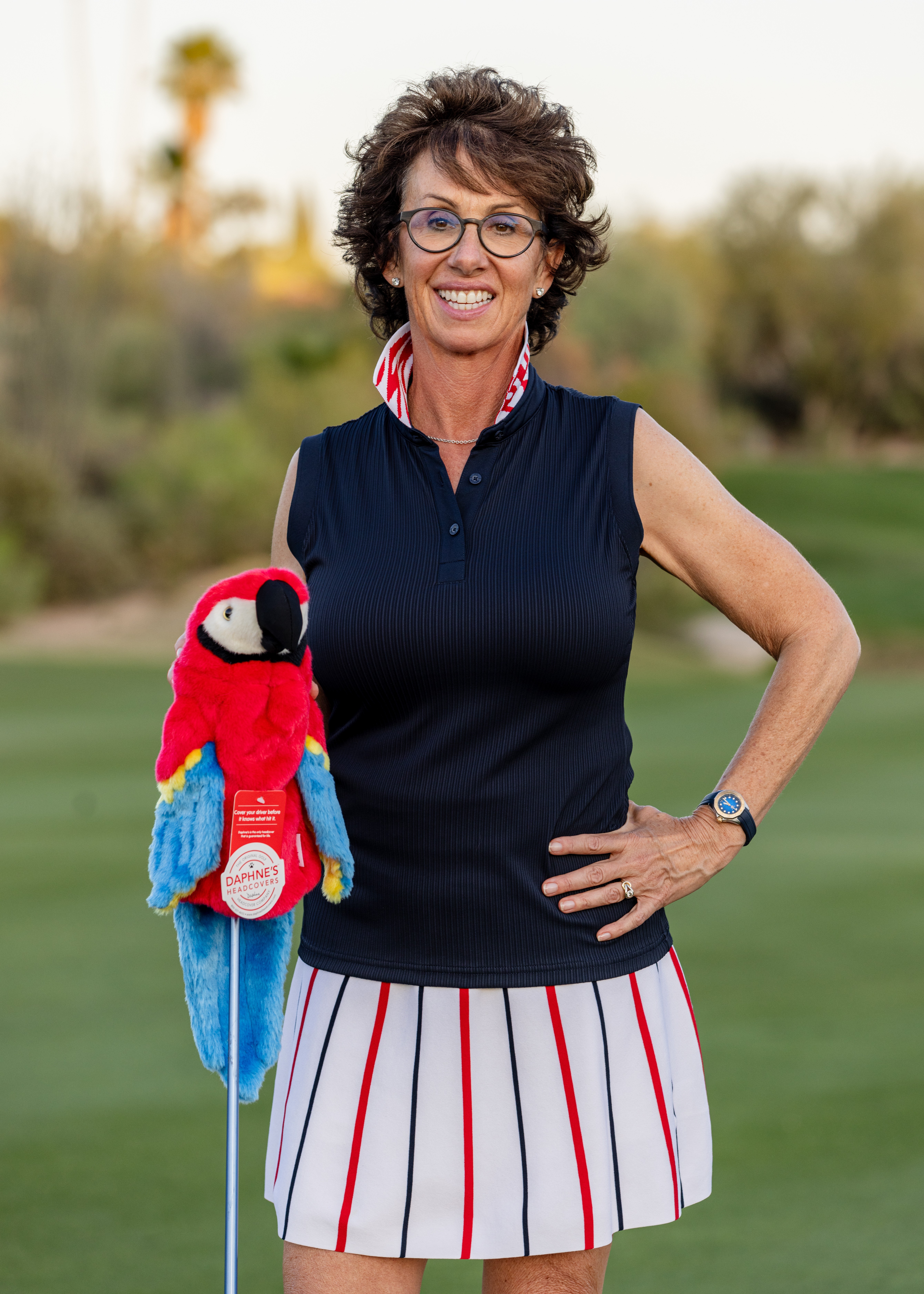Smiling woman with glasses holding a colorful stuffed toucan, standing outdoors on a golf course, wearing a navy top and striped skirt.