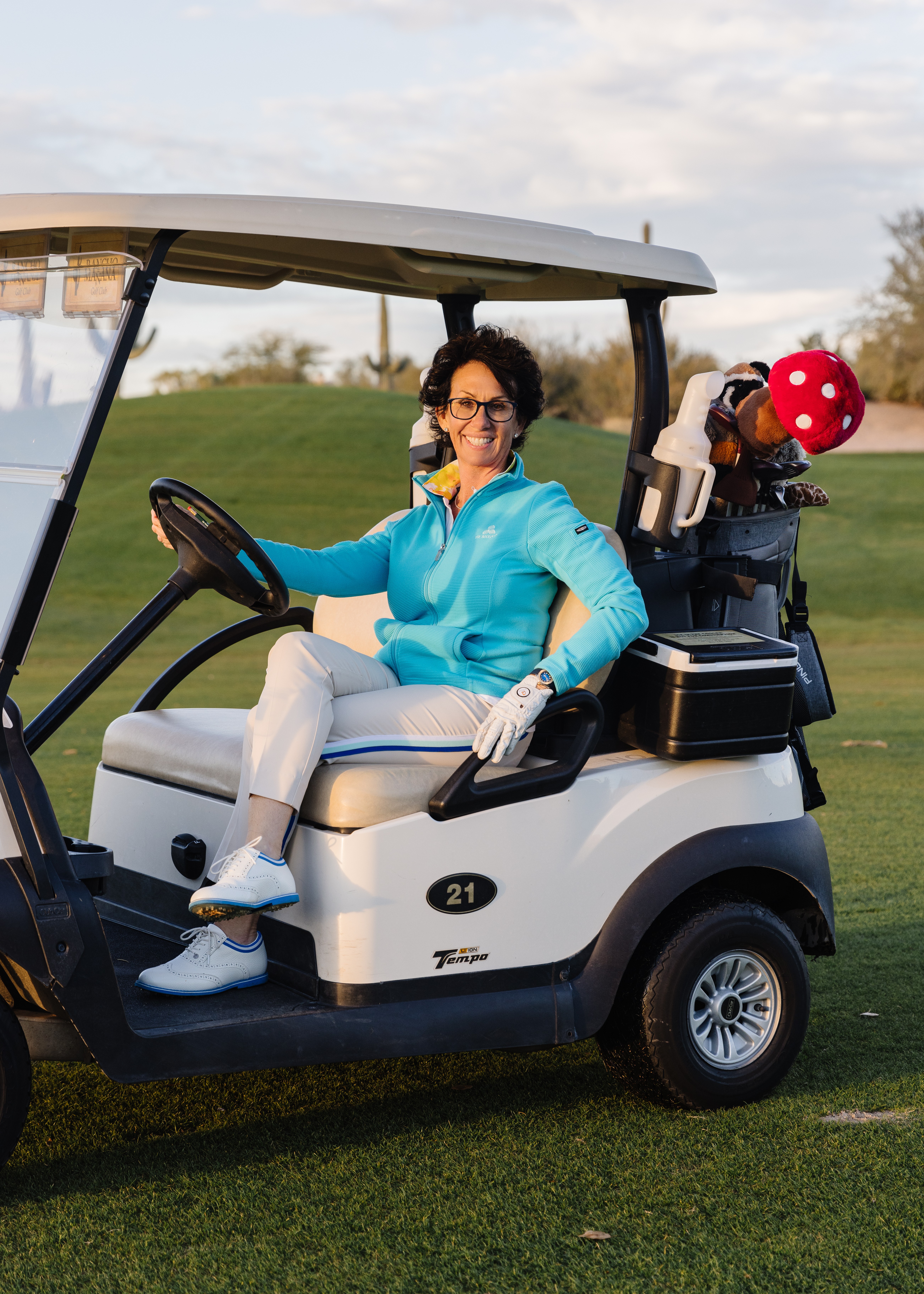 Woman sitting in a golf cart on a golf course, smiling, with golf clubs and a red polka dot hat in the back, outdoors.