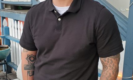 Man smiling, wearing black shirt and gray pants, standing outdoors near a blue structure.