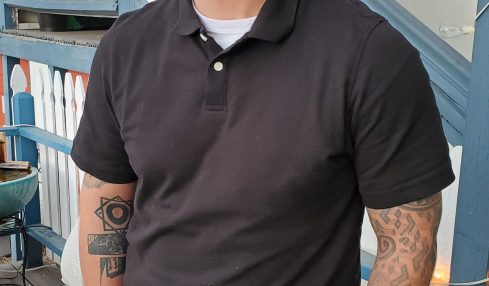 Man smiling, wearing black shirt and gray pants, standing outdoors near a blue structure.