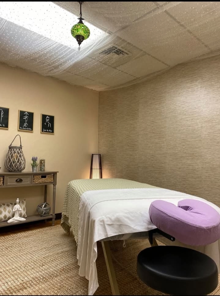 Room with massage table, side table, wall art, and a purple pillow, illuminated by ceiling and floor lamps.