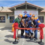 Group of people at grand opening with balloons and ribbon outside a building, smiling and holding scissors, children in front.