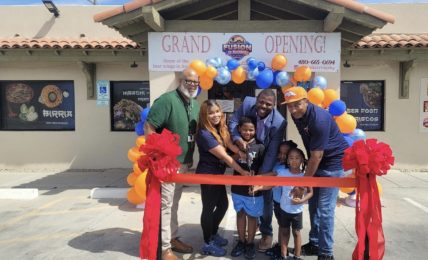 Group of people at grand opening with balloons and ribbon outside a building, smiling and holding scissors, children in front.