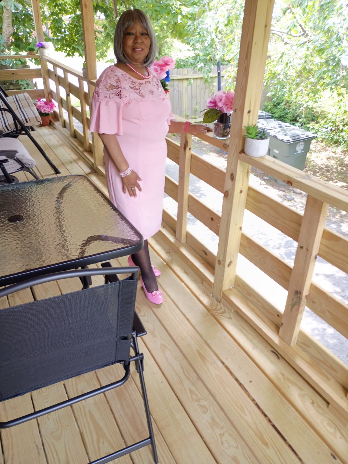 Woman in pink dress standing on wooden porch with flowers and trees in background.