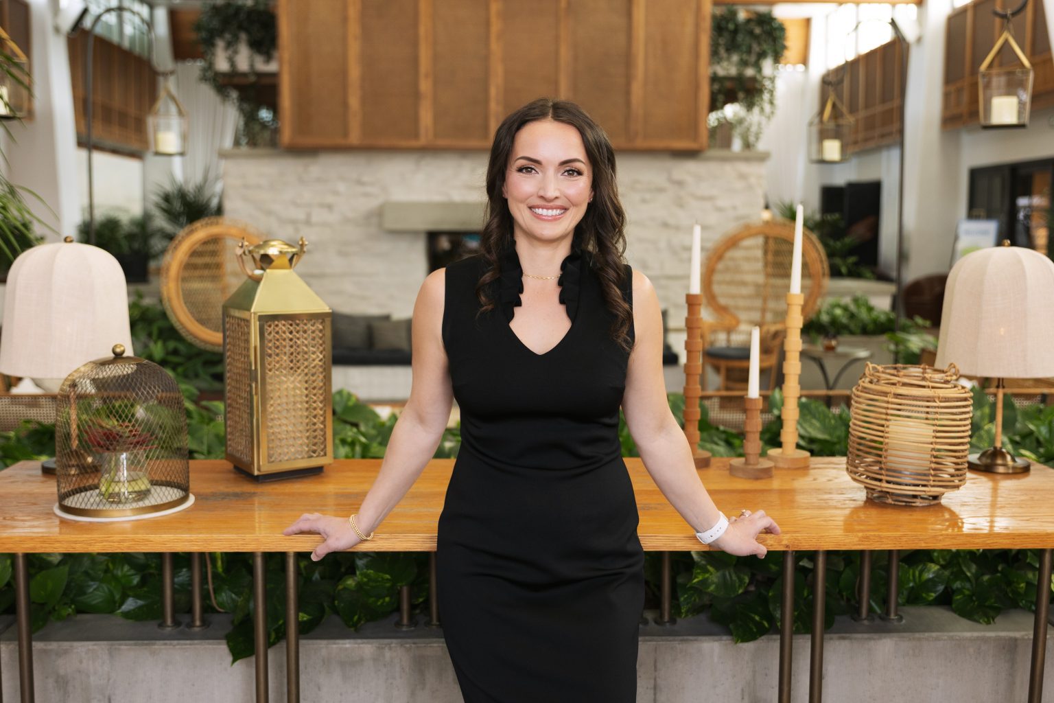 Smiling woman in black dress standing behind a wooden table in a decorated indoor space.