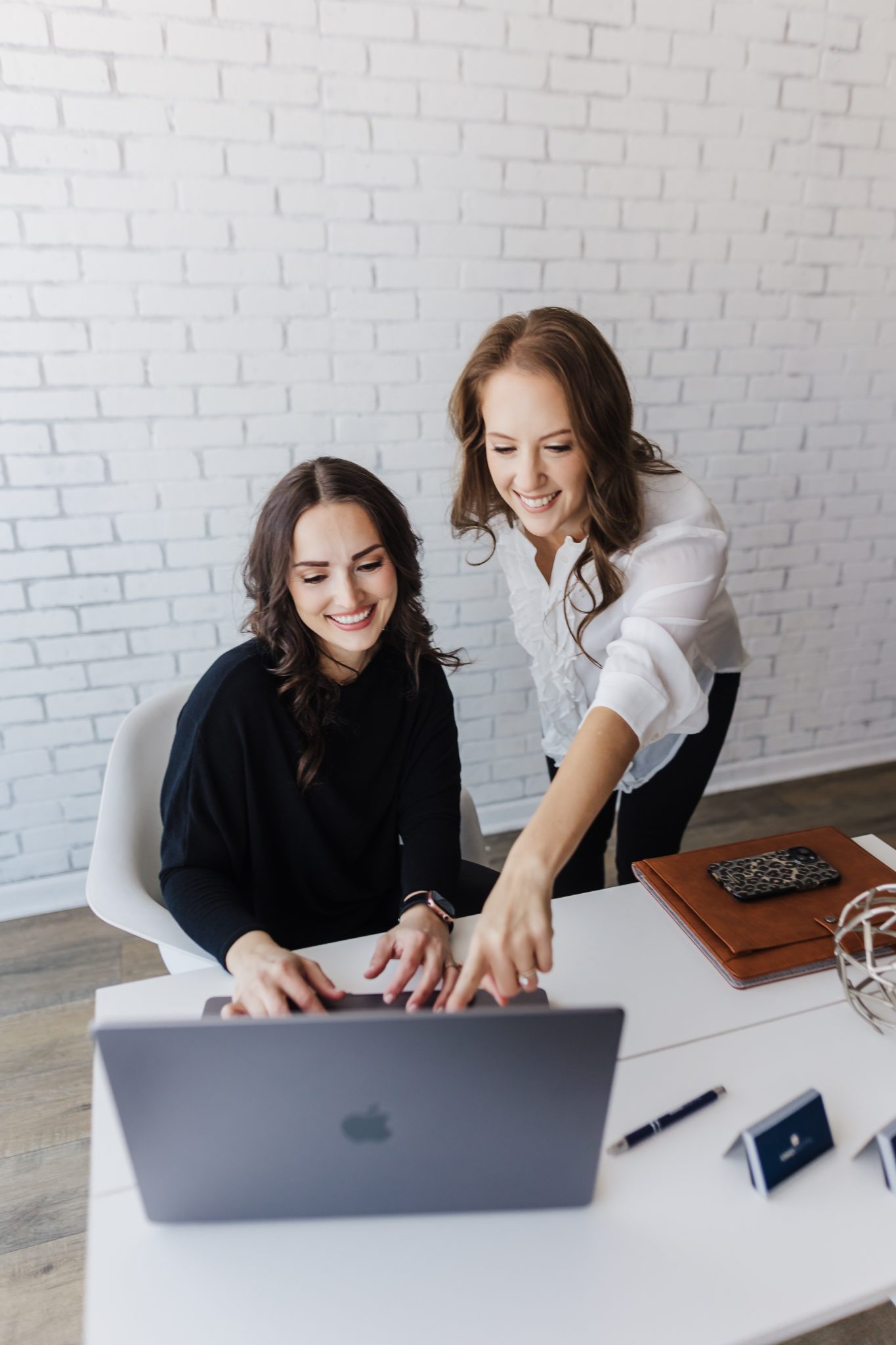 Two women smiling and working together at a desk with a laptop, against a white brick wall.