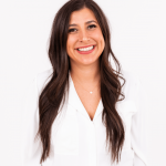Young woman with long dark hair smiling, wearing a white blazer against a plain white background.
