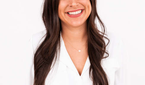 Young woman with long dark hair smiling, wearing a white blazer against a plain white background.
