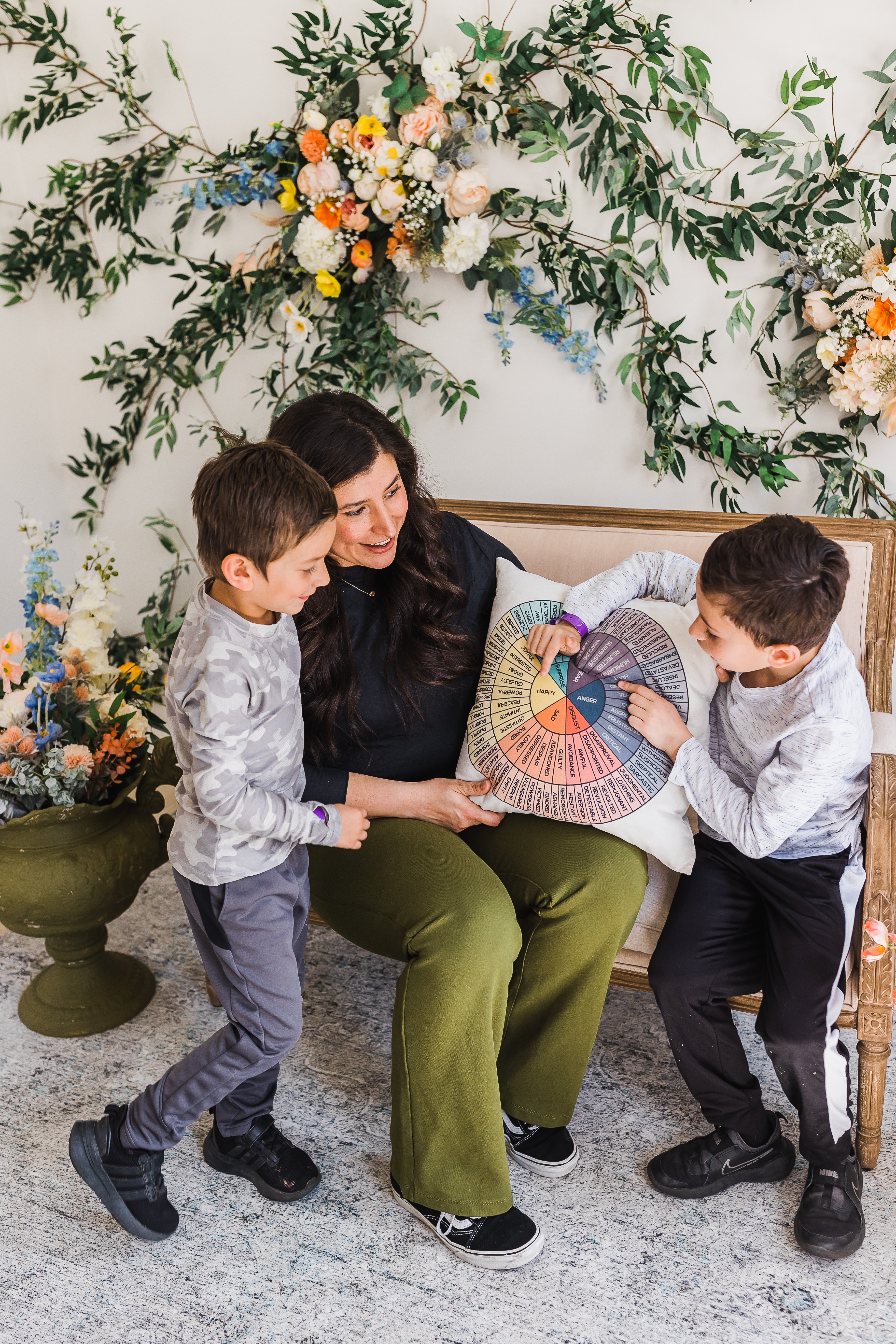 Woman sitting on a sofa with two boys, holding a colorful circular object, surrounded by floral decorations.